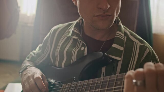 Tilt up shot of focused Caucasian young man with septum nose piercing strumming guitar while relaxing in living room