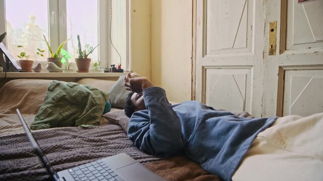 Handheld shot of tired Black freelancer throwing baseball while lying in bed after work on laptop in apartment