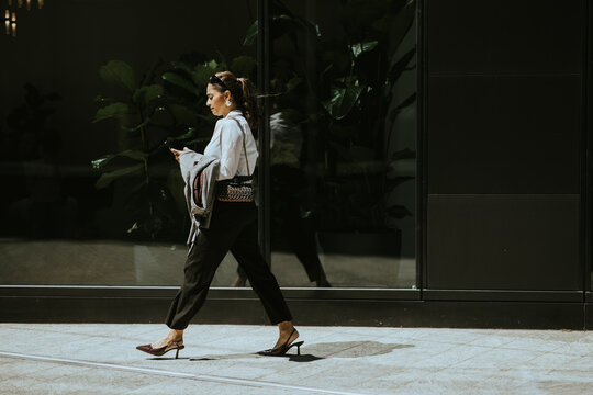 Woman walks in city while using phone near large glass building after midday