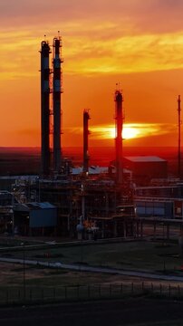 Cinematic aerial view of a petrochemical plant at sunset. Industrial towers, pipelines, and refinery structures stand against a glowing orange sky as warm sunlight reflects across the facility