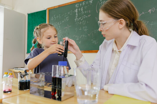 Two female students in a science classroom conduct an experiment using test tubes and liquid, with a chalkboard full of math equations in the background.