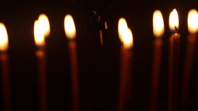 A metal cross, which is held in the hand, approaches a candle among the dense vertical rows of burning church candles in a dim interior, touching the flame,