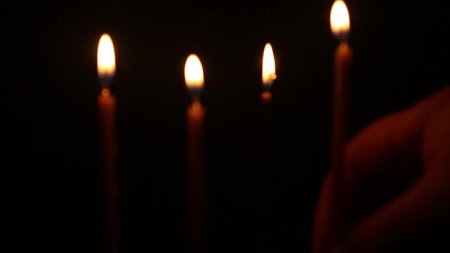 Three church candles burn on reflective dark surface, then a hand enters from below holding lit candle and ignites the fourth, all four flames glow steadily together. no sharpness