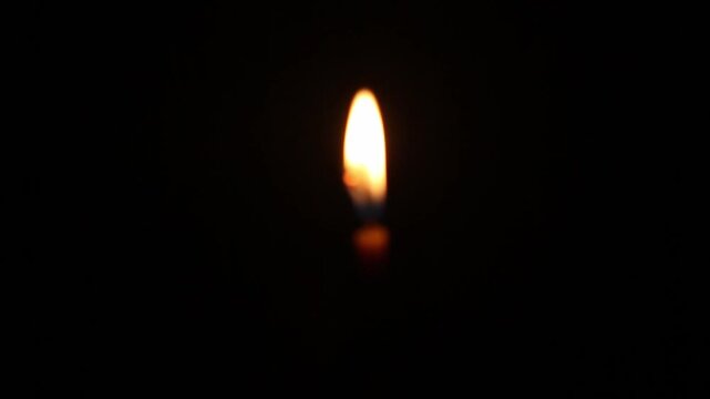 Single tall church candle burns steadily against pure black background, its golden flame gently flickering and slowly swaying, casting warm amber glow in total darkness. no sharpness