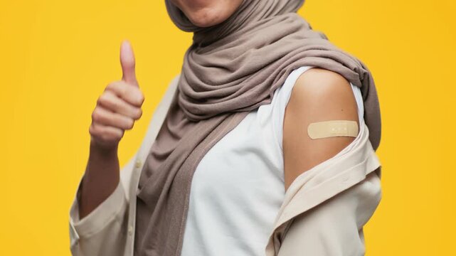 A woman with a hijab shows her arm with a bandage after getting a vaccine. The background is bright yellow. The scene captures a moment of health and wellness awareness.