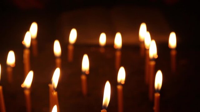 Many church candles burn with bright flames of warm golden tones, filling with sacred light. A man's hand flips through the pages of a book.