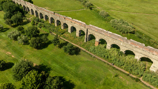 Aerial view of the Parco degli Acquedotti in Rome, Italy. Panorama of the well-preserved stone arches of a Roman aqueduct. The ancient structure cuts through vibrant green fields and scattered trees.