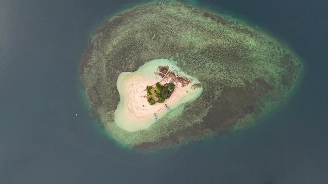 Small tropical island with a sandy beach and dense green vegetation is surrounded by shallow coral reefs and deep blue sea.