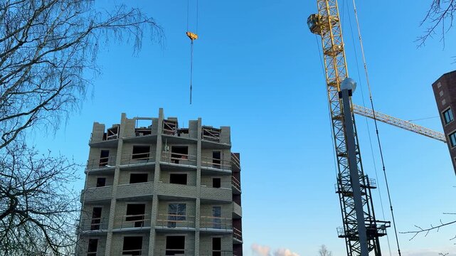 Construction crane above brick building, tower crane hoisting materials beside multistory brick shell under clear blue sky, bare