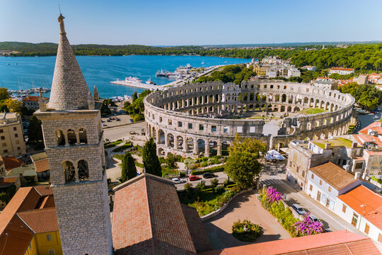Aerial panoramic view of Pula's Roman amphitheatre surrounded by red-roofed buildings, a tall church bell tower and the Adriatic harbor. Sunlit coastal townscape conveying history, Mediterranean charm