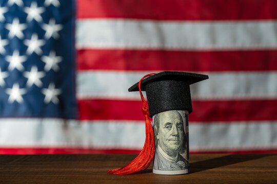 Rolled one hundred US dollar bill wearing black graduation mortarboard hat with a red tassel. Blurred American flag background. Concept for tuition fees or student loans or US higher education cost