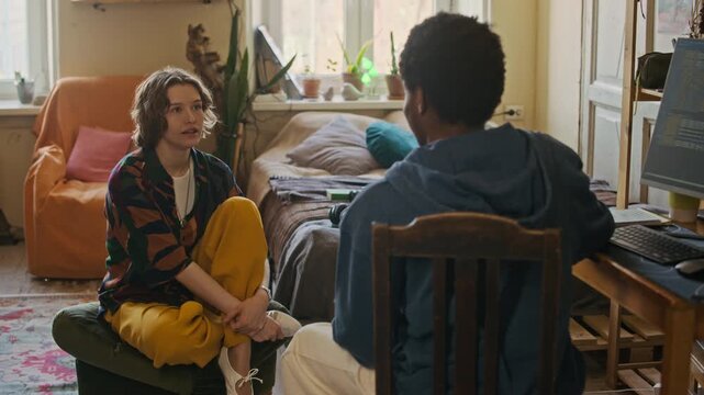 Medium long handheld shot of two diverse gen Z friends sitting in front of each other while communicating in shared apartment, young African American freelancer coding on computer remotely