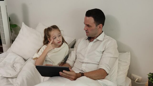 A father and daughter sitting on a bed, focused on a tablet screen together in a cozy, relaxed environment.
