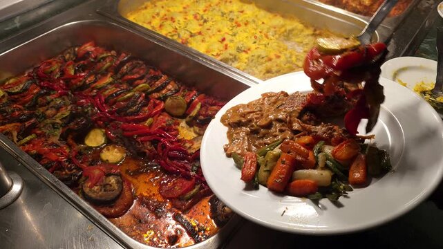 A close-up video of a hotel buffet featuring a large steel chafing dish filled with roasted vegetable ratatouille &mdash; eggplant, red bell pepper, and zucchini cooked in tomato sauce. A man's hand uses a 