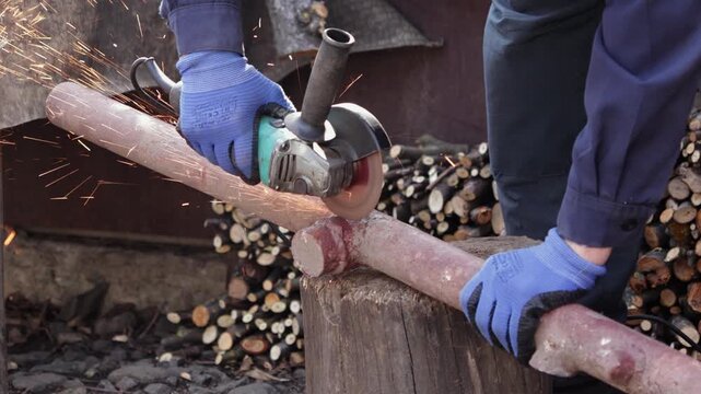 A dedicated worker uses an angle grinder to cut a metal pipe, creating bright sparks during heavy industrial repair tasks in a workshop environment with a wood logs background scene
