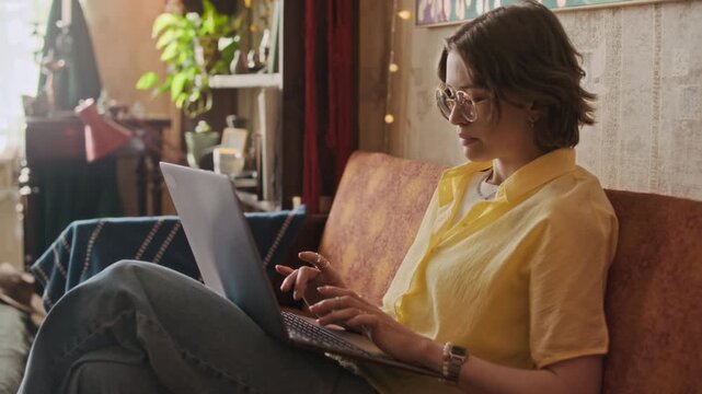 Medium long shot of gen Z female Caucasian freelancer sitting on sofa typing on keyboard of laptop in rented apartment