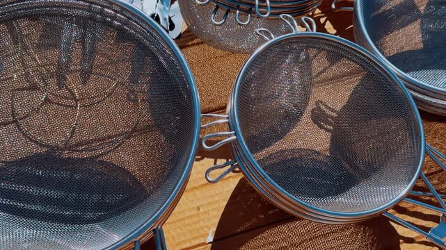 Colander flea market counter close-up showing metallic mesh bowls on sunlit wooden tabletop, assortment of different diameters