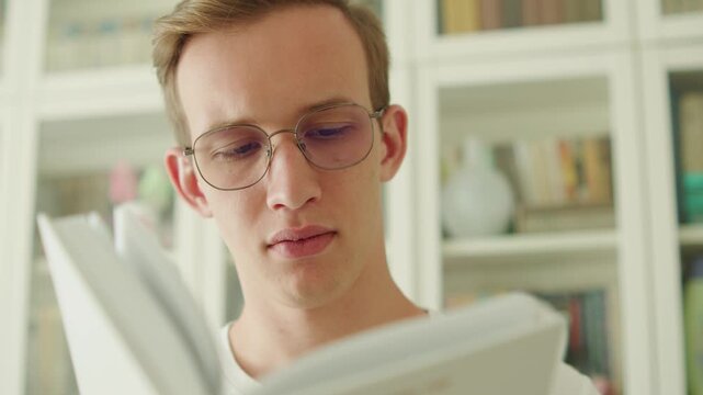 Young man with glasses reading a book in a library. Intelligent teen studying in a library. University student in eyeglasses reading a paperback book in a bookstore. Portrait of a young adult flipping