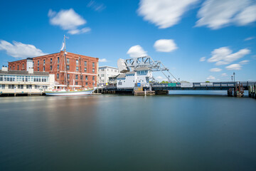 Fototapeta premium Mystic River Bascule Bridge long exposure, a daytime view showing silky smooth water and motion-blurred clouds over the historic drawbridge and docked schooner in Mystic, Connecticut.