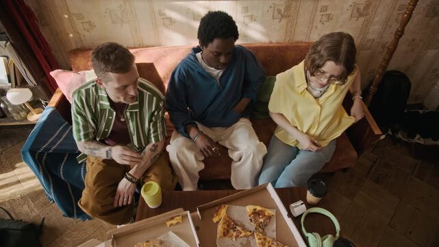 High angle shot of interethnic young roommates checking receipt after eating pizza and splitting bill while sitting on sofa in rented apartment
