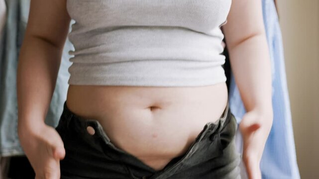 An overweight woman struggles to button up her skinny green jeans after gaining weight, demonstrating the everyday challenges associated with plus-size clothing. High quality 4k footage