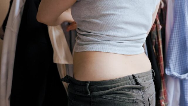 4K video from behind of an overweight woman facing a wardrobe problem, unable to zip up her tight jeans due to weight gain, illustrating plus-size lifestyle annoyance. High quality footage