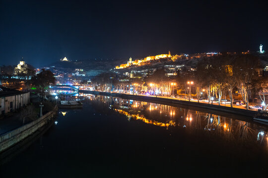 Nighttime cityscape reflected in a calm river, illuminated by streetlights and buildings, creating a peaceful urban atmosphere with glowing reflections and serene water surface.