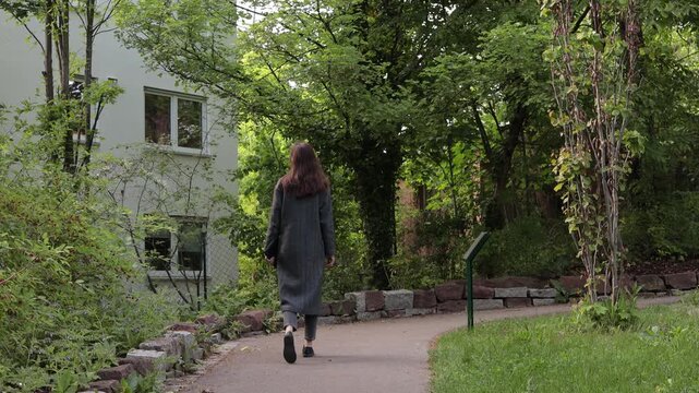Woman walking on a path beside a building surrounded by lush green trees and plants