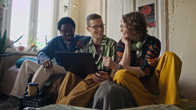 Long handheld shot of three diverse young friends watching film on laptop while spending time together in shared apartment