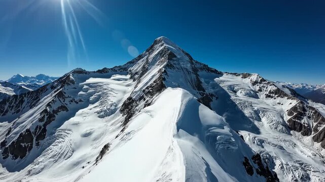 Aerial view of a sharp snow-covered mountain peak and glacier under a clear blue sky with bright sunlight and lens flare, showing detailed icy ridges and high alpine terrain.