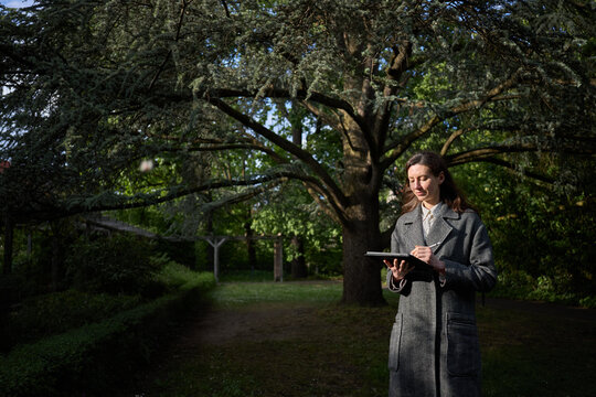 Woman in a coat using a tablet computer outdoors under a large tree