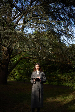 Woman in a long coat using a tablet under a large tree in a park