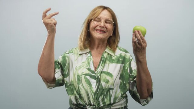 Woman middle age senior with blonde hair and glasses wearing leaf patterned shirt holds a green apple in one hand and points finger with the other hand in studio backdrop; joy wellness.