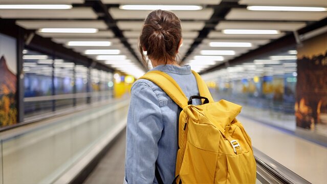 Woman with a yellow backpack standing on a moving walkway in a modern airport terminal, carrying luggage for solo adventure, experiencing travel, and starting a new journey