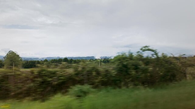 Rapid side view from a moving vehicle showing the verdant fields and lush forests of the rural Asturian region under a cloudy sky, evoking a sense of fast travel and motion