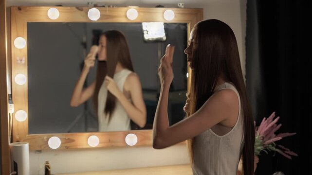 A woman with long brown hair combing her hair in a dressing room while looking in the mirror