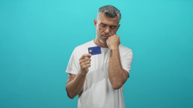 Man holding creditcard and resting hand on cheek while looking down in studio against a turquoise seamless wall; financial worry uncertainty.