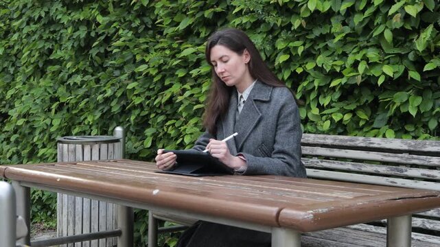 Woman working on tablet at outdoor table with green hedge background