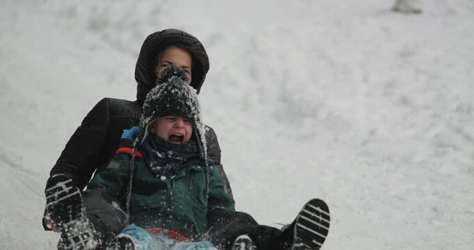 Mother and little son on saucer sled sliding fast on mountain slope, spreading the snow around, winter joy in slow motion