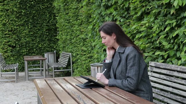 Woman working on tablet at outdoor cafe table with lush green hedge background