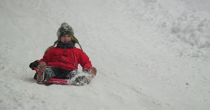 Little boy on saucer sled sliding fast on mountain slope, spreading the snow around, winter joy in slow motion