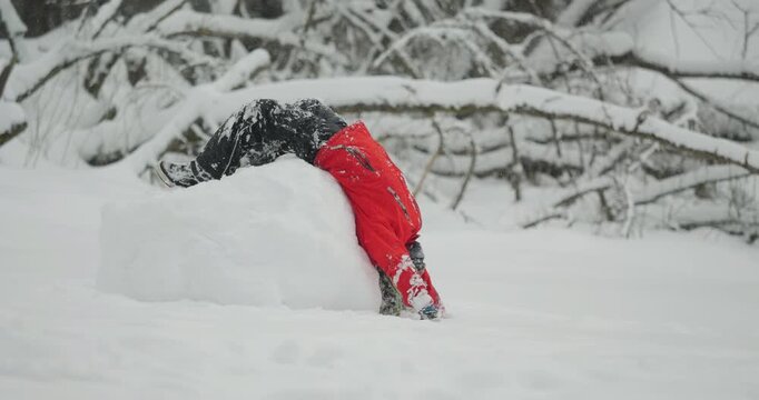 Child in a red jacket playfully leaning backward over the top of a giant snowball, then rolls upside down
