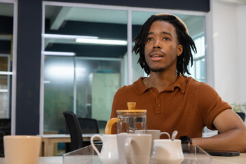 Fototapeta premium African American man sitting at office table in rust polo, French press, copy space