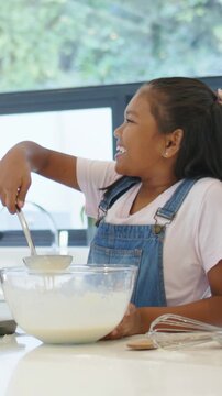 Vertical video: Steadying child preparing batter to bake at counter, in overalls holding glass bowl