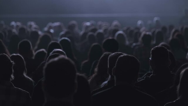Silent dark cinema crowd watching film, rows of silhouetted heads seen from back, moody atmosphere, focus on audience and anticipation.