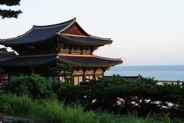 Obraz premium Buddhist temple Hyangiram near sea at sunrise. Traditional Korean architecture with layered roof. Green plants surrounds old religious building.