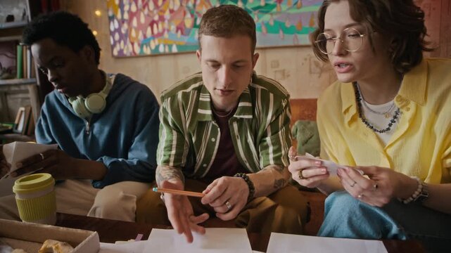 Handheld shot of three young roommates calculating past due utility bills and receipts before paying while sitting at table with pizza in shared apartment