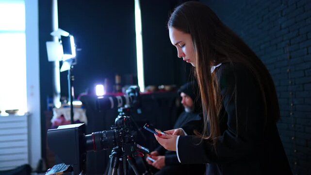 Studio Assistant Checking Smartphone Beside Video Camera. Assistant checks smartphone beside tripod camera, with LED lights and black brick wall in background.