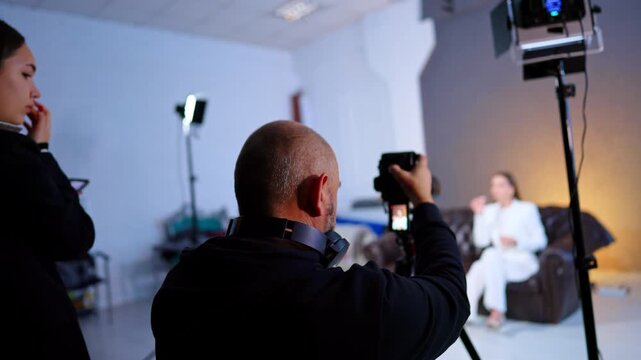 Camera Operator Filming Interview In Photo Studio. Camera operator films seated woman on couch while assistant monitors setup under studio lights.