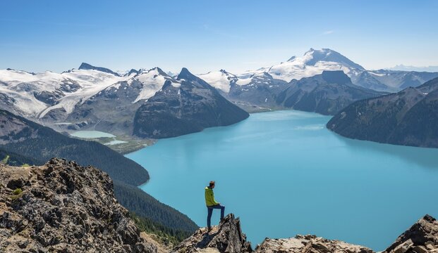 Young man standing on a rock, looking into the distance, view of mountains and glacier with turquoise blue lake Garibaldi Lake, peaks Panorama Ridge, Guard Mountain and Deception Peak, Garibaldi Provincial Park, British Columbia, Canada